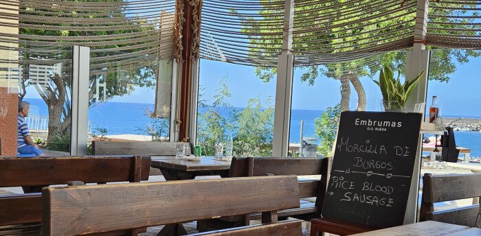 Utomhusrestaurang med utsikt över havet, trätak, meny för blodkorv, lediga bord och stolar, klar himmel.