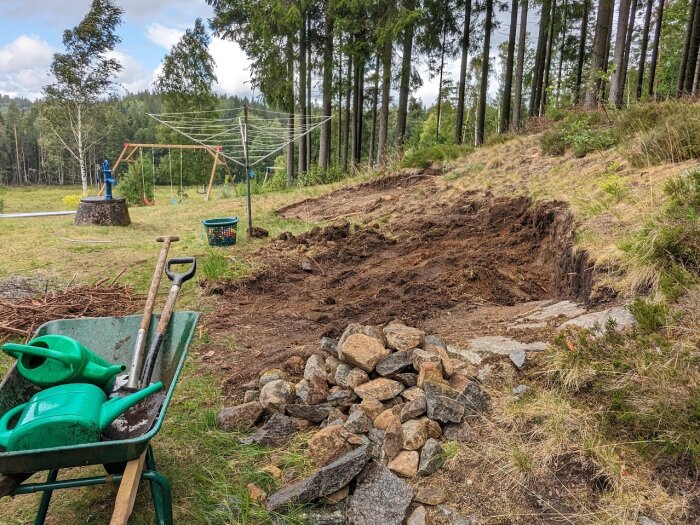 Utgrävningsarbete i grönområde med skottkärra, spadar, vattningskanna och stenar; lekplats i bakgrunden.