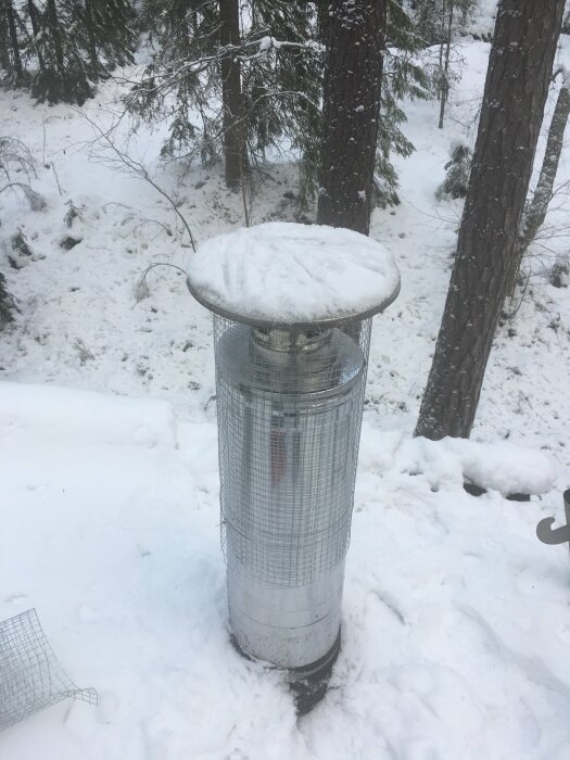 En metallcylinder i snö med snötäckt topp, i en skogsmiljö.