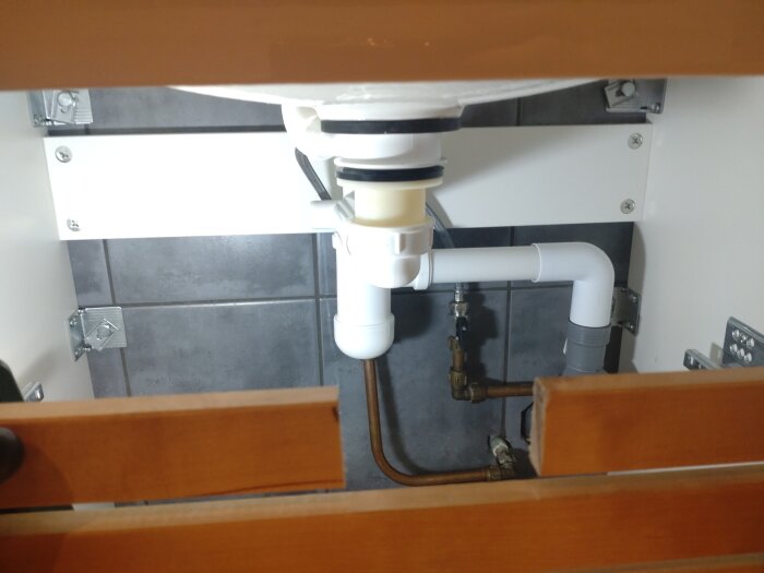 VVS under diskbänk: avloppsrör, vattenledningar, vinkelventiler och grå kakelvägg.