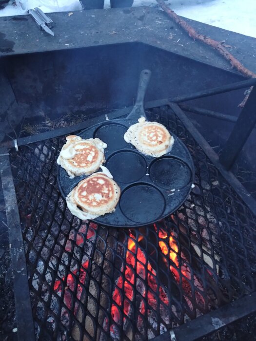 Pannkakor steks på gjutjärnspanna över glödande kol i utomhusgrill vid vintertid.