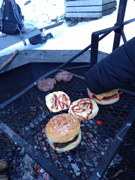 Grillning utomhus på vintern, hamburgare med bröd och sås, snötäckt mark, ryggsäck, grillgaller över glödande kol.