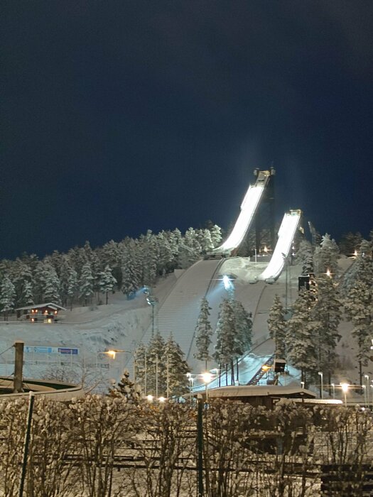 Kvällsvy av upplysta skidbackar med snö och skog, med en hoppbacke i centrum.