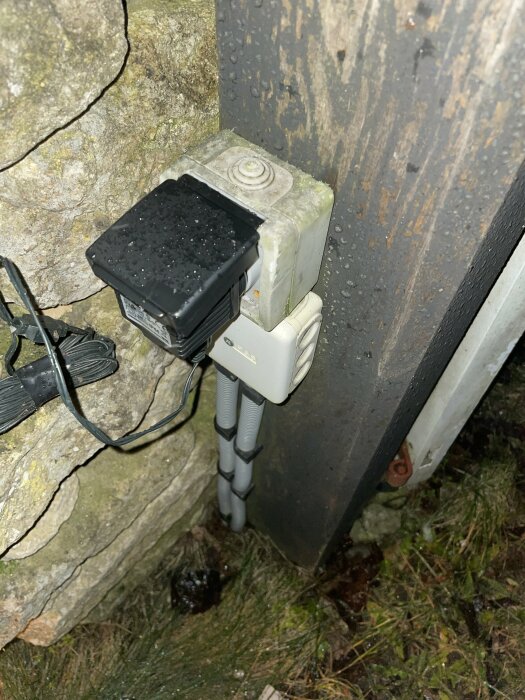 En väderbeständig kamera fastsatt på en trästolpe nära en stenmur och elektriska kopplingar, möjligen för övervakning.