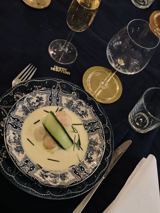 Elegant middagsdukning med sopptallrik, dekorativt mönster, champagneglas, mörk duk, "Happy New Year"-dekoration.
