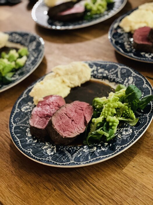 Ett måltidsarrangemang med skivat kött, romanesco broccoli och puré på ett mönstrat blått fat.