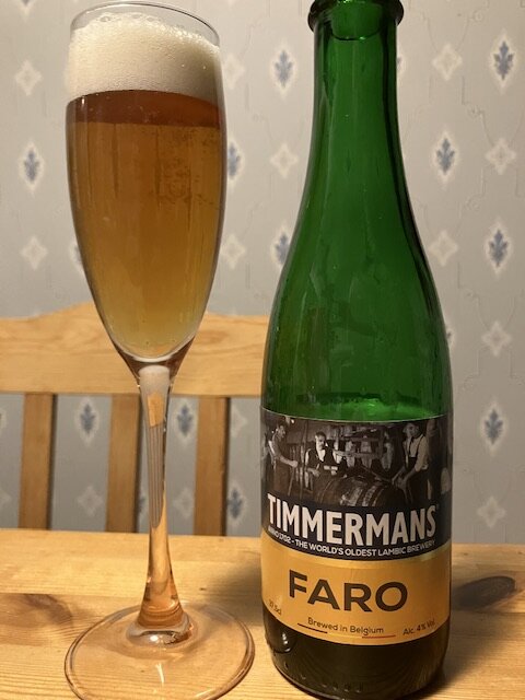 En grön flaska Timmermans Faro-öl bredvid ett fyllt champagneglas på ett träbord.