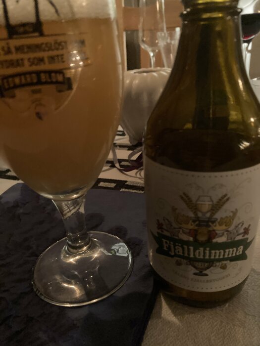 Ölglas fyllt med ljus dryck, tom ölflaska, "Fjälldimma" etikett, suddig bakgrund, inomhusmiddagsmiljö.
