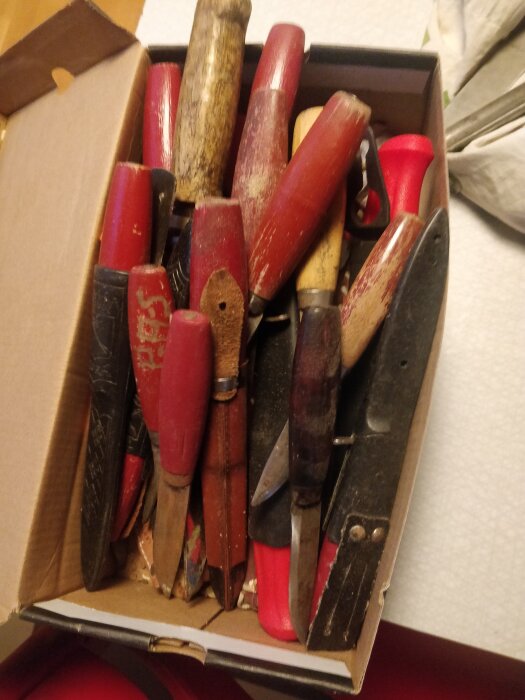 Låda med använda verktyg: tänger, mejslar och knivar med röda och trähandtag.