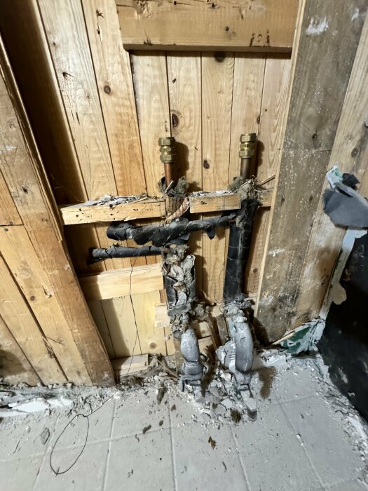 Skadade rör och ventiler i en vägg, omgivna av trä och rörisolering, med synligt byggavfall.