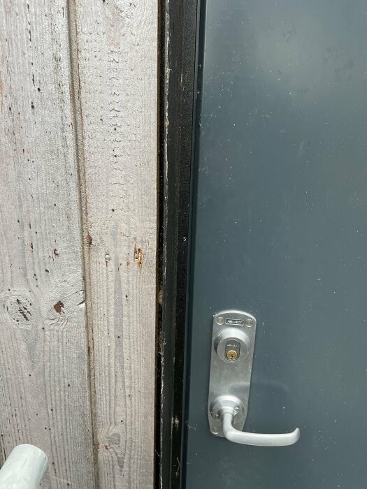 En grå dörr med handtag och låskista, intill träyttervägg, synlig gångjärnsida och dörrtröskel.