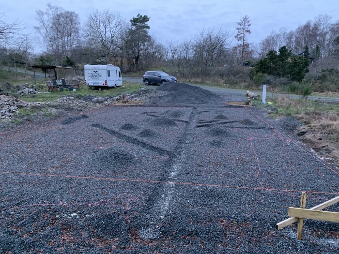 Grusplan med markeringar, husvagn, bil, skogsbryn, och gryning eller skymning. Byggplats eller parkeringsyta under anläggning.