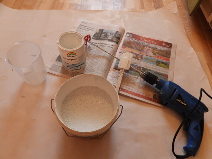 Målningsmaterial på täckt golv: hink, färg, pensel, mätbägare, borrmaskin med visp.