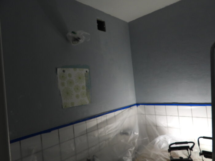 Renovering pågår, grå vägg, blå tejp, oskyddade eluttag, delvis täckt kakel, skyddsplast, oavslutad inredning.