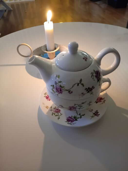 Blommönstrad tekanna och kopp på fat med tändljus på vitt bord.