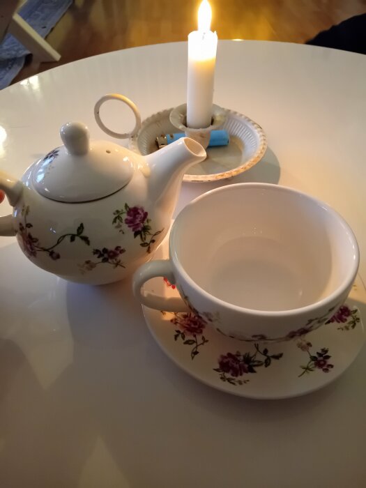 Tekanna och kopp med blommönster, tänd ljus, mysig atmosfär, vit bakgrund, avkopplande te-tid.