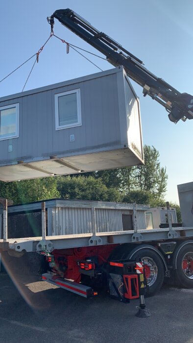 En kran lyfter en byggnadsmodul från en lastbil under en klart blå himmel.