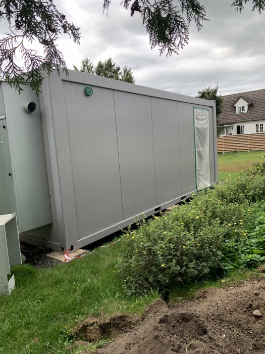 Stor container eller modul, troligen tillfällig, i trädgård, hus i bakgrunden, mulet, icke färdigställd installation.