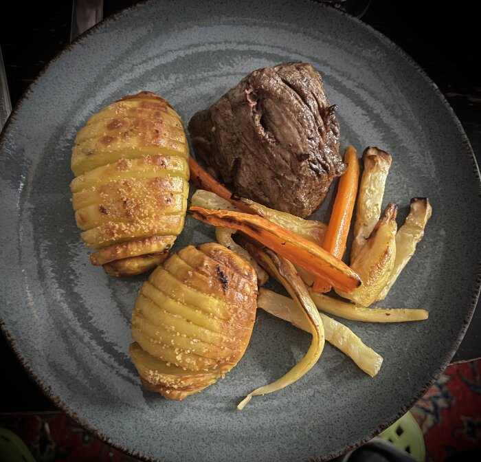 En tallrik med stek, hasselbackspotatisar och rostade morötter och palsternacka. Välkomponerad matpresentation på grå tallrik.