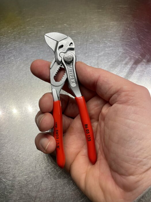 En hand håller en Knipex tång med röda handtag på en metallisk yta.