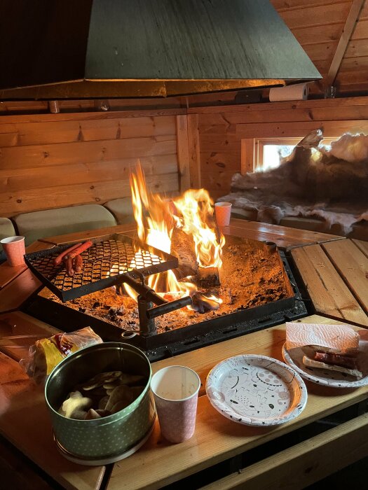 Eld i öppen spis, trähytt, fika med mackor och kakor, mysig atmosfär, träväggar och fönster.