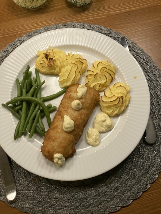 Ett måltidsfat med friterad fiskfilé, gröna bönor, duchessepotatis och klickar av sås, serverat på ett vitt fat.