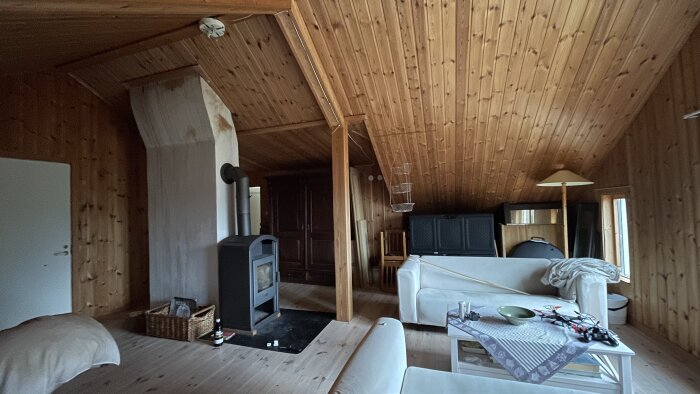 Ett hemtrevligt vardagsrum med träbeklädnad, vedspis, soffor och matbord, inrett i lantlig stil.