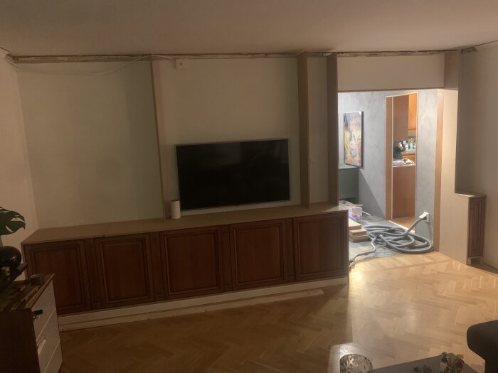 Vardagsrum under renovering, trägolv, hängande TV, skåp, oavslutad vägg och tak, kabel på golvet.