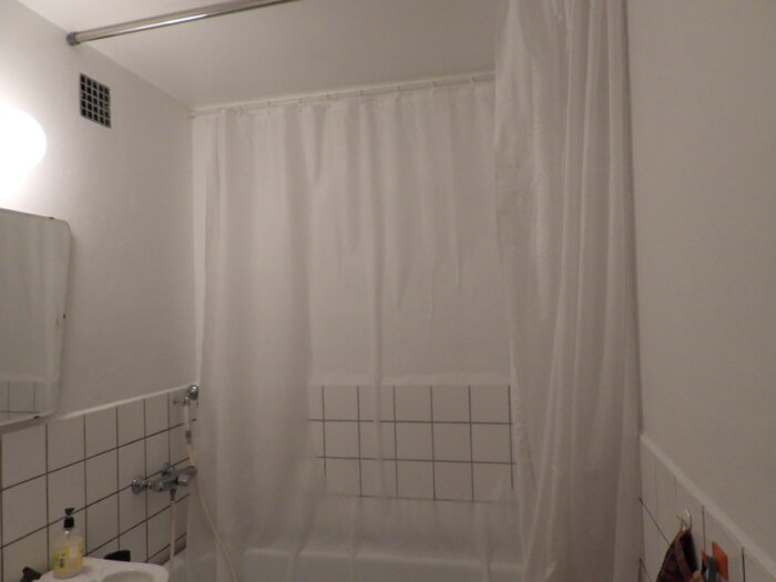 Enkelt badrum med vit duschdraperi, kaklad vägg, dusch, badprodukter och vitmålad vägg.