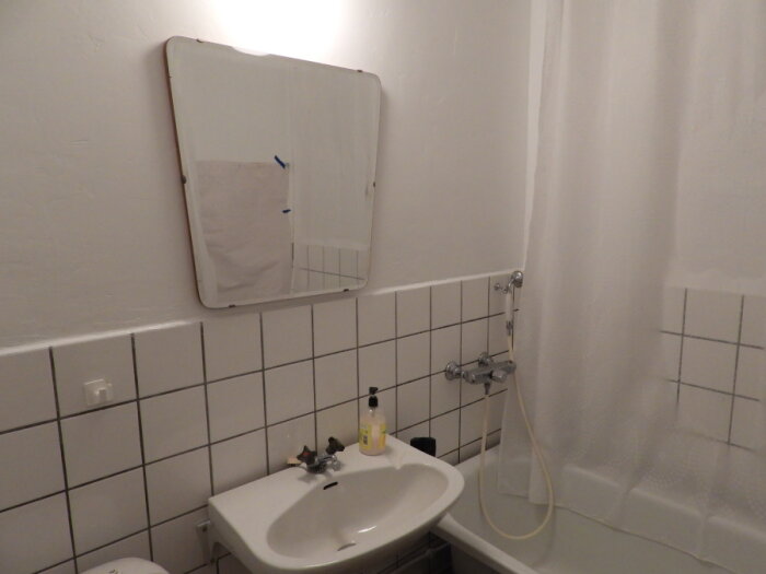 Ett enkelt badrum med spegel, handfat, tvål, och badkar med draperi. Klinkergolv och väggar med vita kakelplattor.