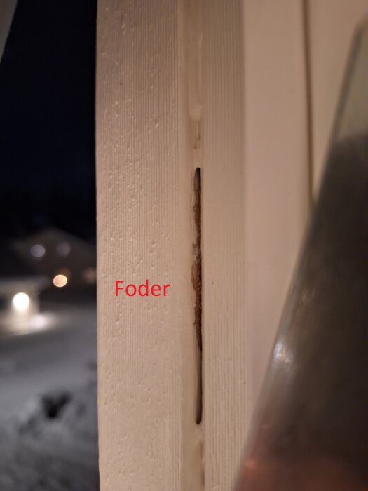 Närbild på skadad vitmålad dörrkarm med spricka, markerad med texten "Foder", natt och snö utomhus.