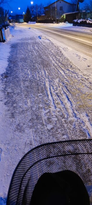 Vintergata, snöigt, belyst, blå timme, kalla färger, fordonsspår, del av en barnvagn i förgrunden, tätbebyggt område.