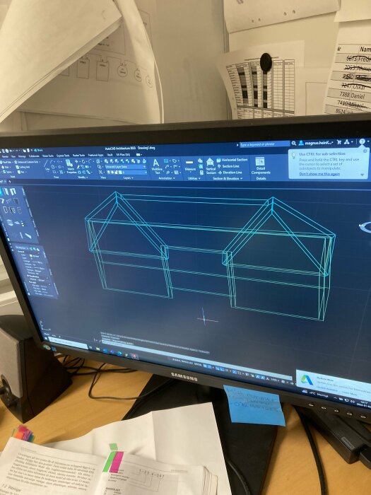 Datorskärm visar CAD-program med ritningar av husstrukturer, kontorsarbetsplats, pappersdokument.