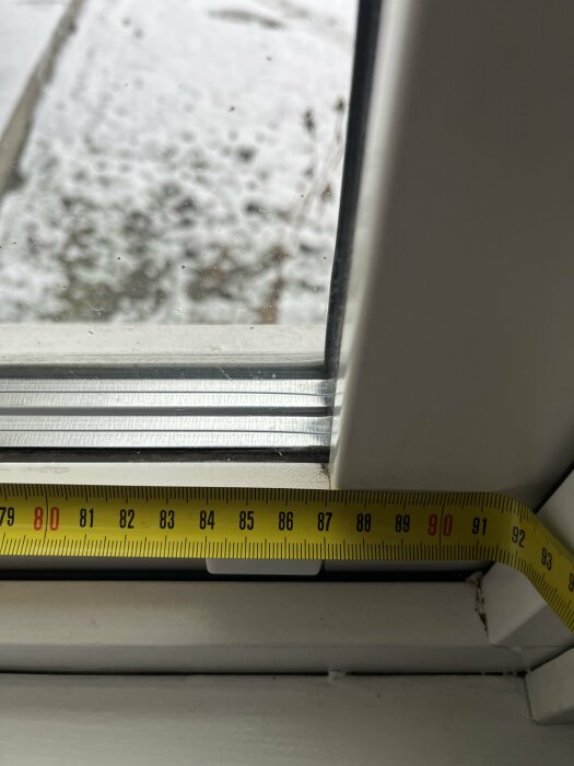 Mätband vid fönster, visar bredd i centimeter, utomhus snöig utsikt suddig i bakgrunden.