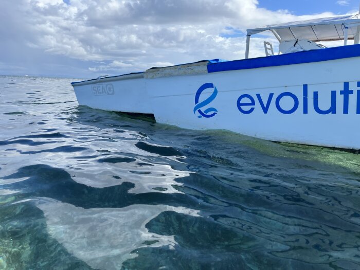 Vit båt med text "evolution" flyter i klart, blått vatten under en molnig himmel.