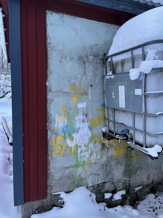 Vintervy: snötäckt vattentank, färggrann graffiti på slitet betongvägg, röd träkarm.