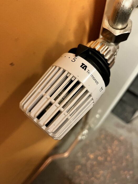 Termostatventil på radiator, inställt värmevärde, vita och svarta detaljer, metallanslutning, brun bakgrund.