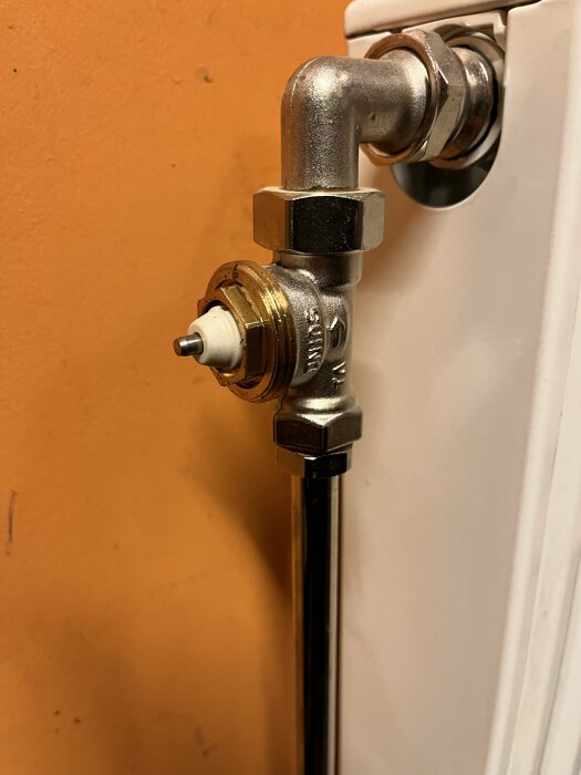 Radiatorventil i första plan, ansluten till värmeelement, mot orange vägg, sidovy, inomhusinstallation.