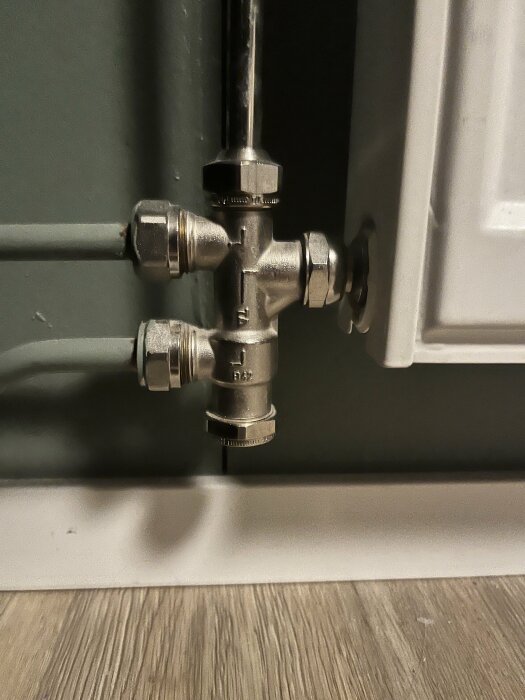 Värmeelementanslutning med rör, avstängningsventil och termostatventil. Grå vägg, vit golvlist och trägolv syns.