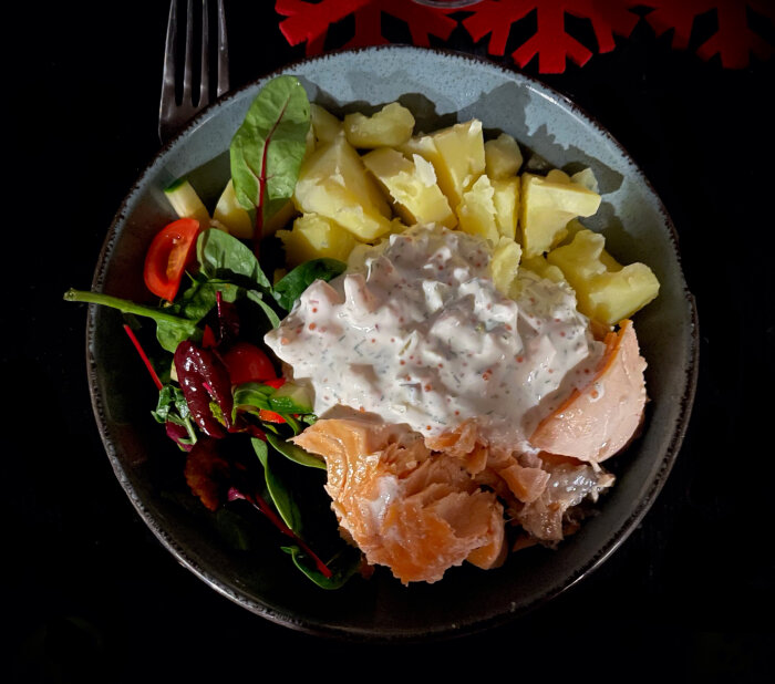 En tallrik med potatis, rökt lax, sallad och gräddfilssås, serverad på en mörk bakgrund.