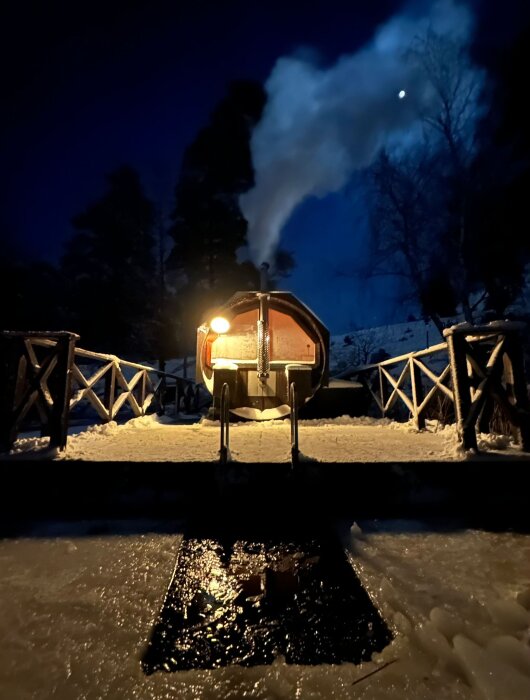Ett upplyst tåg framför rök, mörk himmel, träsnöig plattform, kväll, kall atmosfär, reflektion på is.