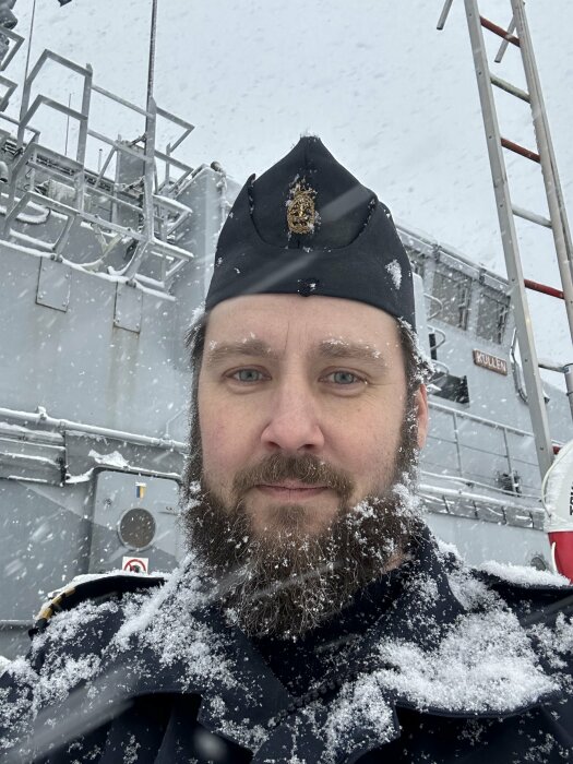 Man i militär uniform med snö i skägget framför industriellt metallstruktur i vintermiljö.
