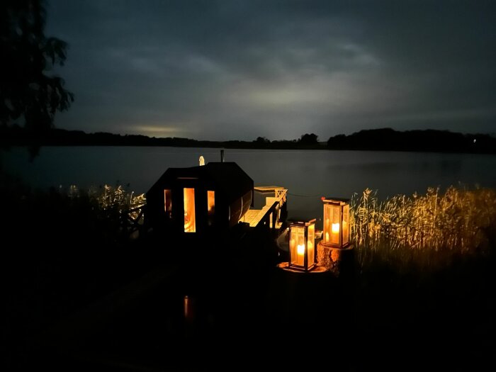 Ett litet hus vid sjön med belysning vid skymning, reflektioner i vattnet, lugn och fredlig stämning.