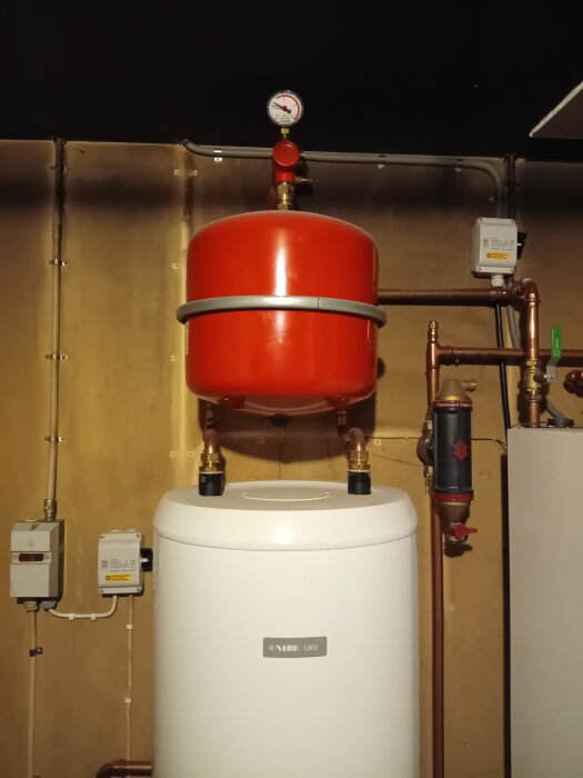Värmesystem med expansionskärl, tryckmätare, vit varmvattenberedare och kopparledningar mot trävägg.