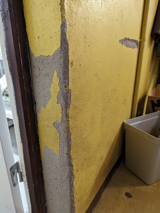 Gul vägg med färg som flagar bort, exponerar underliggande betong, bredvid fönster och dörrpost.