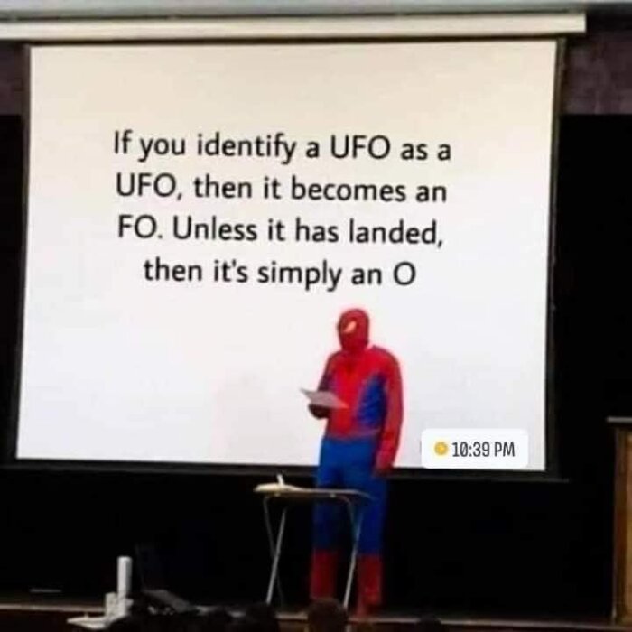 Person utklädd till Spider-Man föreläser, humoristisk UFO-text på skärm, tidstämpel i hörnet.