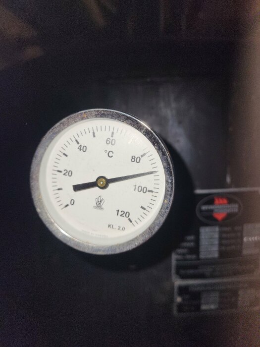 Analog termometer visar cirka 60 grader Celsius, monterad på utrustning med etiketter, industriellt sammanhang.