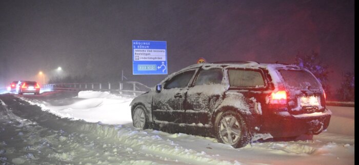 Bil i snöstorm vid väg, med trafikskyltar, natt, nödblinkers, djupt snötäcke, vinter, kallt, nödsituation, dålig sikt.