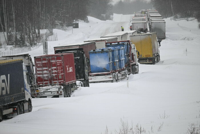 Lastbilar i kö på snöklädd väg, vinterförhållanden, trafikstopp, snöfall syns, personer vid fordon.