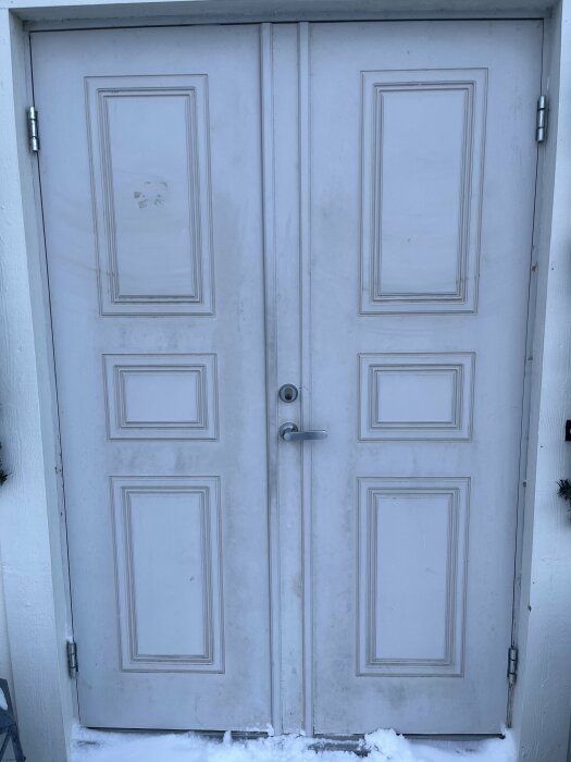 Dubbla vita dörrar med paneler, dörrhandtag, snö på marken, behöver rengöring.
